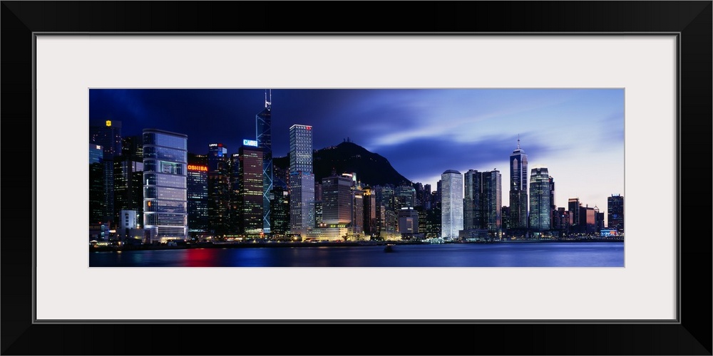 Panoramic photograph displays the bright skyline of a famous city reflecting over a portion of Victoria Harbour.  In the b...