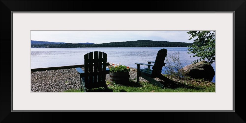 A panoramic photograph of Adirondack chairs arranged with a view of a lake on a bright sunny day.