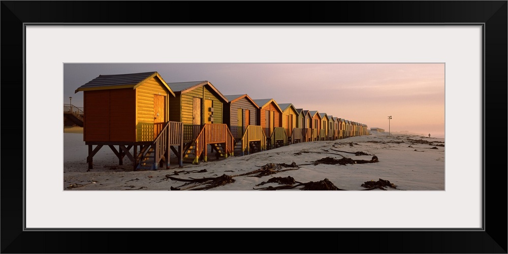 A panoramic photograph of a row of buildings built on the sandy shoreline glowing in the light of sunset.