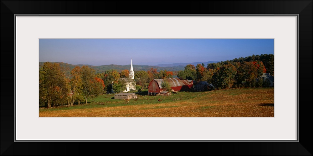 This panoramic photograph is of a barn and a church surrounded by autumn trees with large hills in the distance.