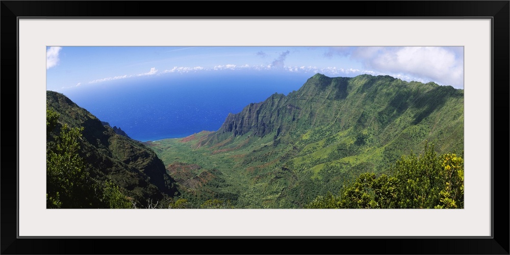 Horizontal photograph on a large wall hanging of a green cliff that overlooks the blue sea, beneath a sky with few small c...