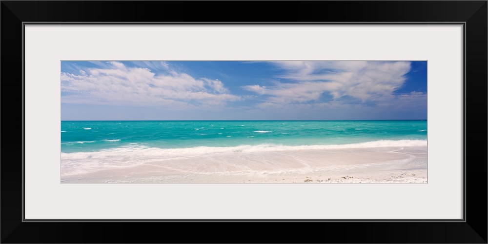 Panoramic photograph of calm ocean with surf and sand in the foreground and cloudy sky above.