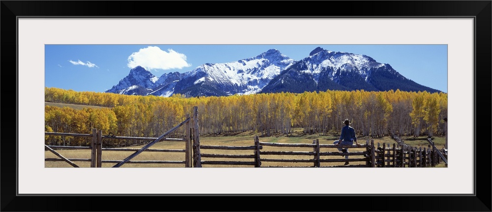 Large panoramic image of a cowboy sitting a top a pasture fence looking towards the fall colored trees and mountains.