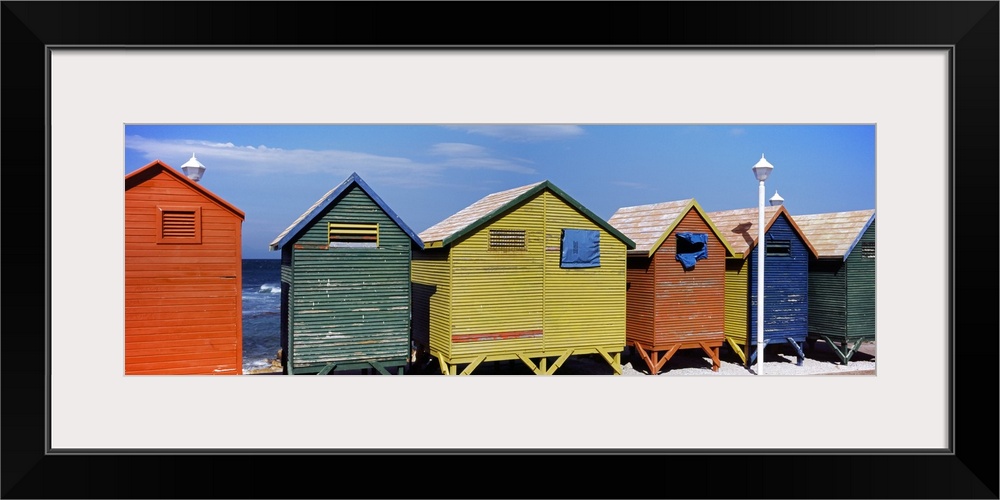 Panoramic photograph taken of multi-colored huts that line the beach with the ocean viewed in the distance.