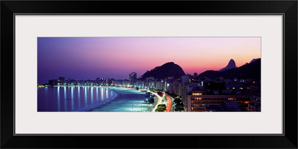 Panoramic, giant photograph of buildings lit along Copacabana Beach at night, mountains in the background against a vivid ...