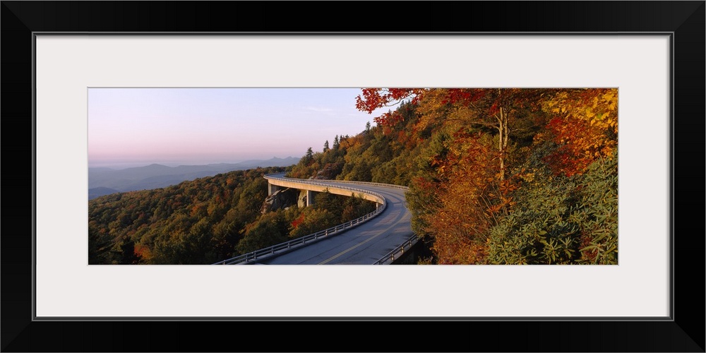 Panoramic photograph of winding mountain road with tree tops below it and mountain silhouette in the distance.