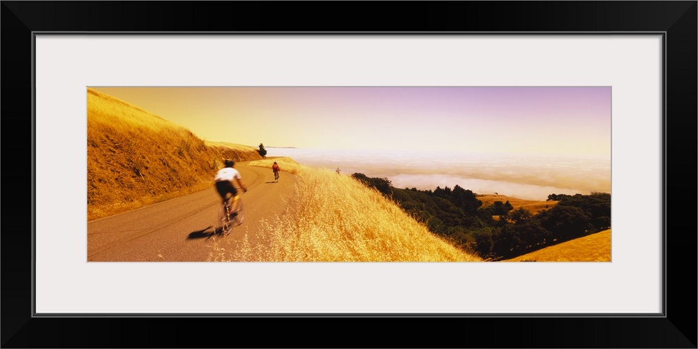 Long canvas of two people riding bikes on a paved path curving on top of a cliff overlooking a forest and with water in th...