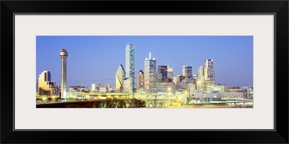 Giant, landscape photograph of a highway road leading toward the lit up Dallas skyline against a blue sky, at dusk.