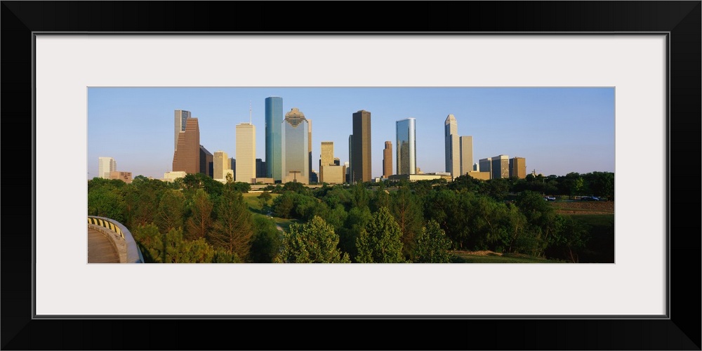 Skyscrapers in the Houston skyline are photographed from a distance with large trees in the foreground.