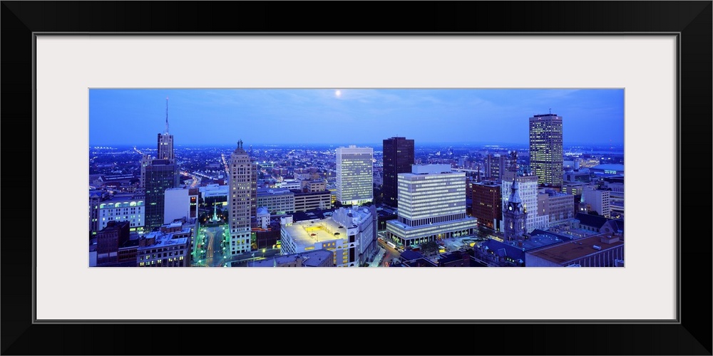 This panoramic wall art is an aerial photograph of the city skyline at dusk with moon shining down on the city.