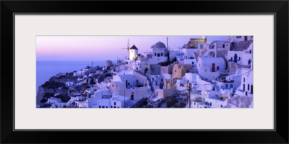 Wide angle photograph of various buildings on a sloping hillside near the coast, in Santorini, Greece, at sunset.