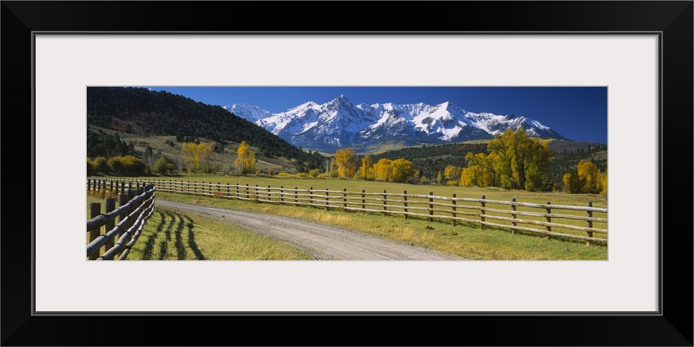 Panoramic photograph of dirt path lined by a wooden gate with snow covered mountains in the distance under a clear sky.