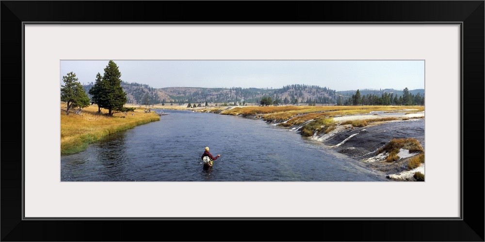 Wide angle shot of a person standing in thigh high water fishing with a beautiful view of park terrain in front of them.