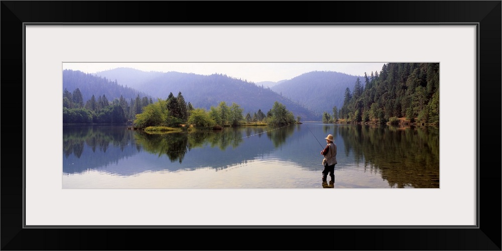 A fisherman stands in shallow water with his line out. There is a beautiful view of trees and large hills in the distance.