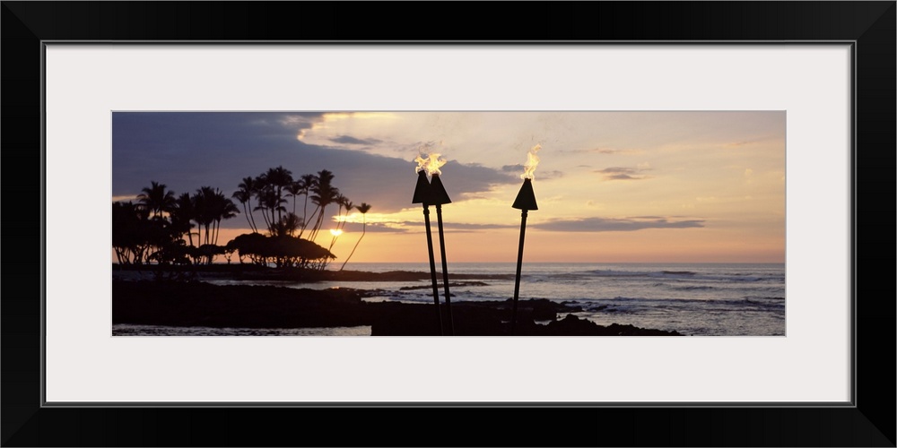 Panoramic photograph of silhouetted palm trees and land reaching into dark waters beneath the setting sun.  Several lit to...