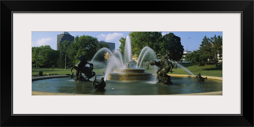 A large fountain with various statues in it is photographed in panoramic view with a park shown in the background.