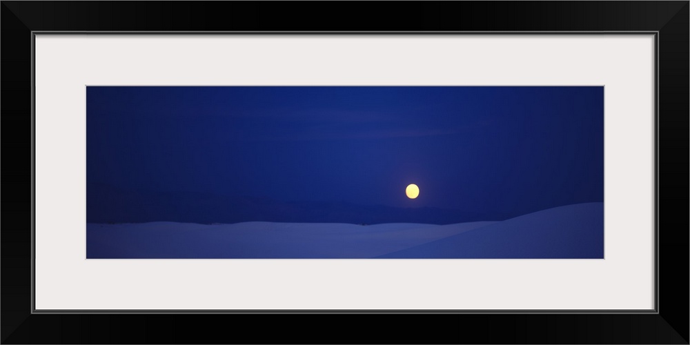 Panoramic photograph taken of a full moon against a deep blue sky and sitting above white sand that is shaded blue.