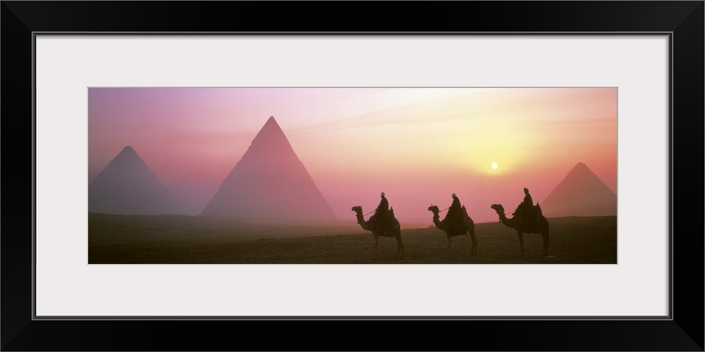 Panoramic photograph of three camel riders at dusk with pyramids in the background.  The sun is setting and is masked my t...
