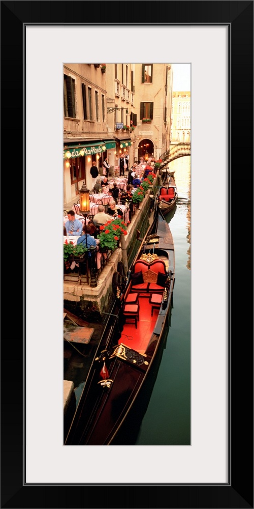 Vertical panoramic photograph boats on canal waterway lined with street cafes and buildings.