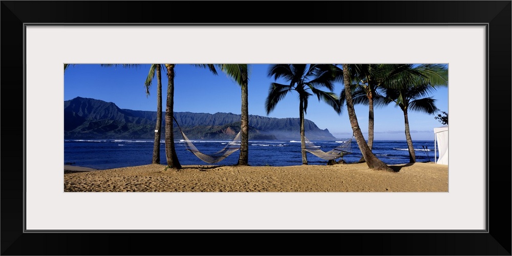 Panoramic print of two hammocks swaying between palm trees along the ocean with mountains in the distance.