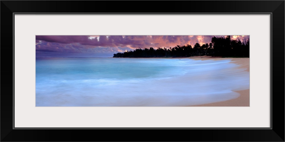 A time lapsed photograph of waves lapping against a sandy tropical beach with the silhouetted of a jungle in the distance.