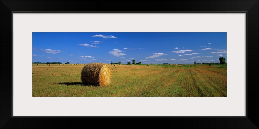 A large panoramic piece of a single bale of hay in a wide open field with an almost cloudless blue sky above.