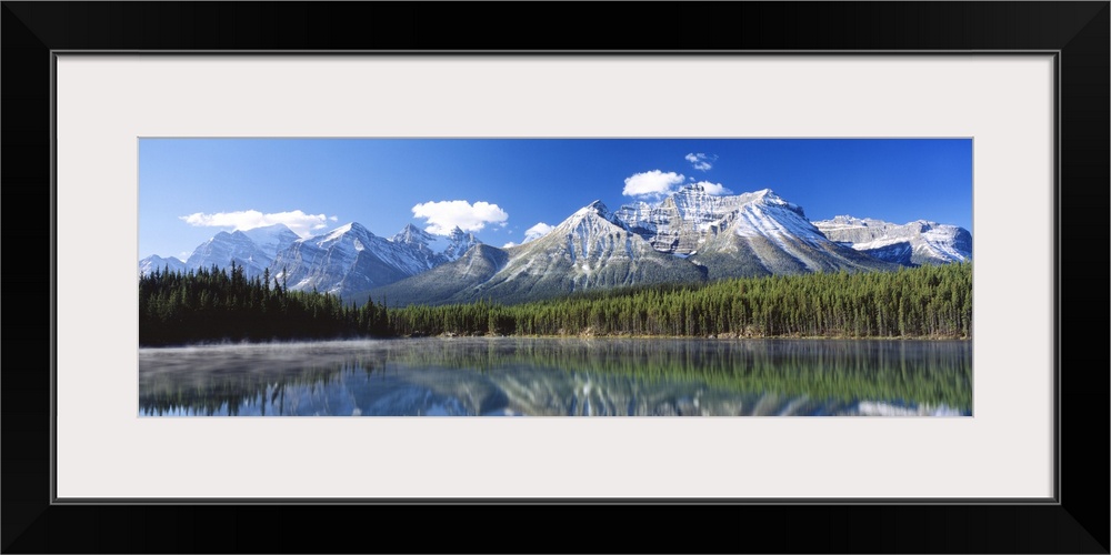 Large panoramic canvas of big snowy mountains with a dense forest beneath it reflected in the water.