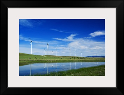 Herd of cattle grazing beneath row of wind farm turbines, reflection in pond water, Montana