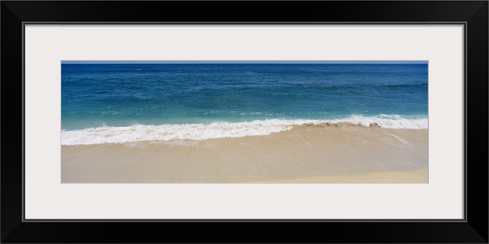 Long photograph of waves crashing in to the sandy beach as blue water stretches for miles in the distance.