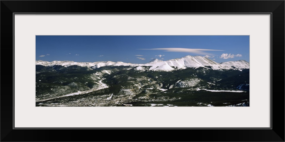 This wide angle photograph is an aerial shot of a snow covered mountain range with vast land and a town in front of them.