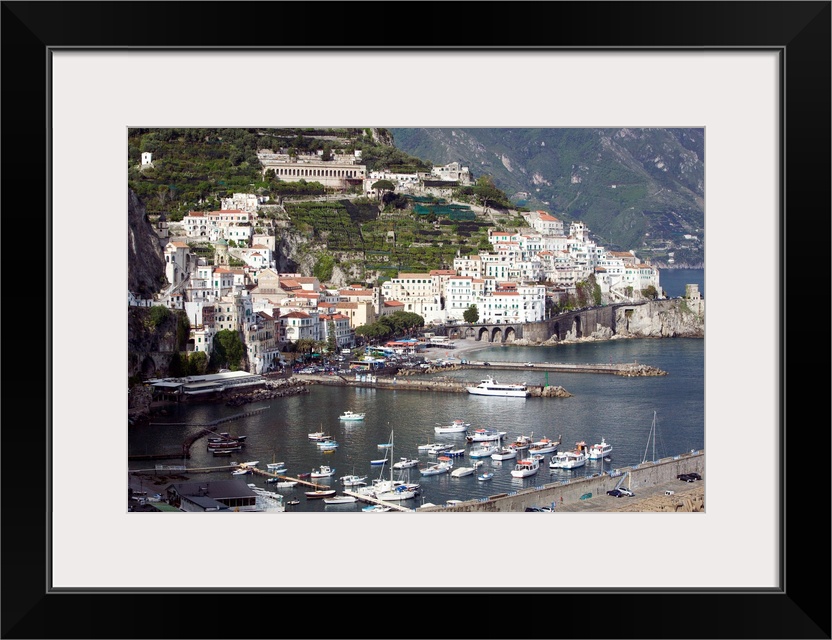 Photograph of busy port town with boats coming in to dock.  Part of the water's edge is made of mountains filled with buil...