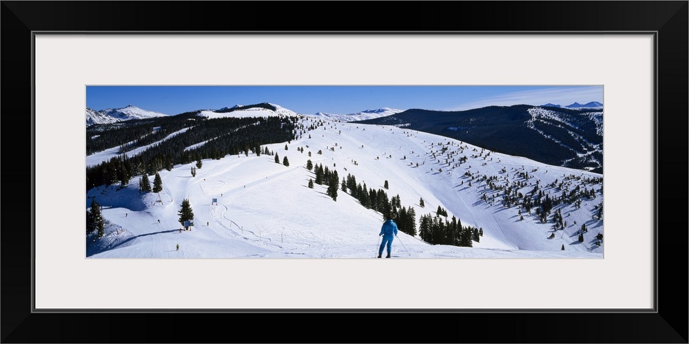 A panoramic photograph of a skier departing down a snow covered slope at the top of a mountain.
