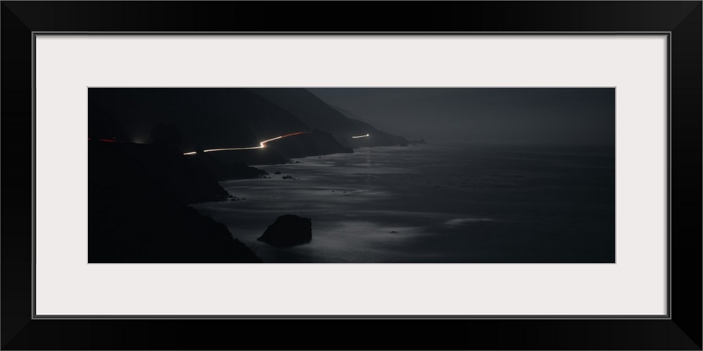 Panoramic photograph of road near coastline lit up with moving headlights at night.