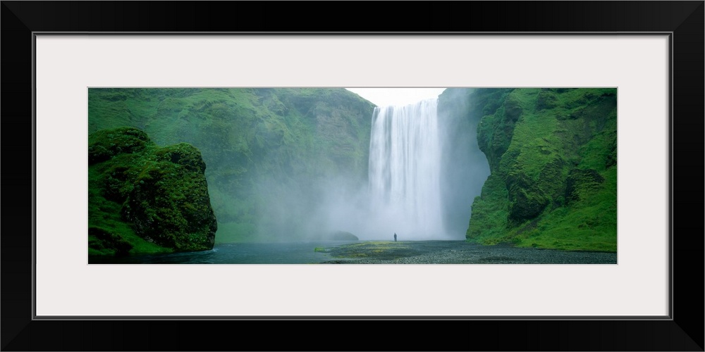 Panoramic photo of a wide waterfall spilling over a cliff into the water below with a man standing on the land in front.