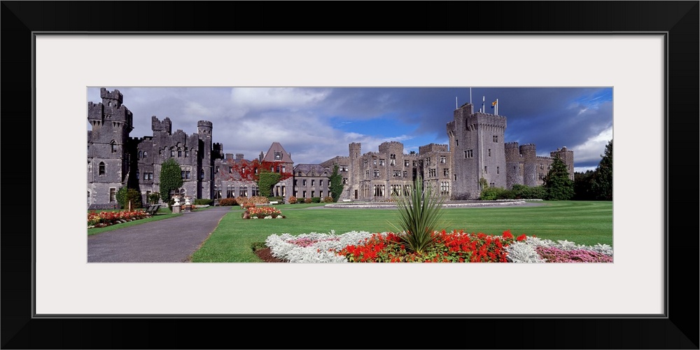 Panoramic photograph taken of an immense castle that has lush green grass in the front yard filled with planted flowers.