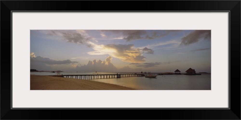 Panoramic photograph of pier stretching into ocean from the beach under a cloudy sky at dusk.