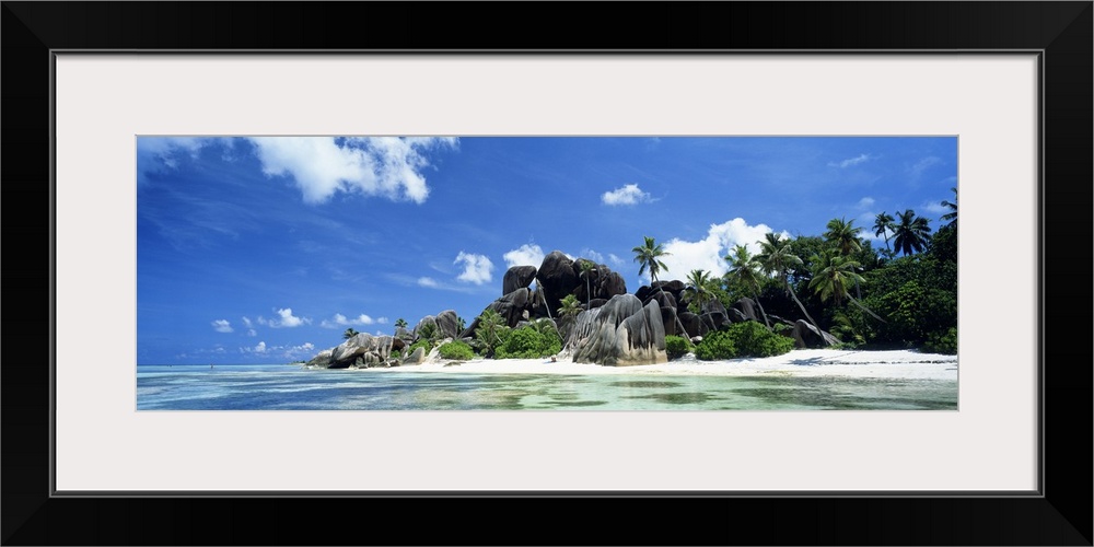 A panoramic photograph of a tropical beach lined with large boulders and palm trees taken on a sunny day.