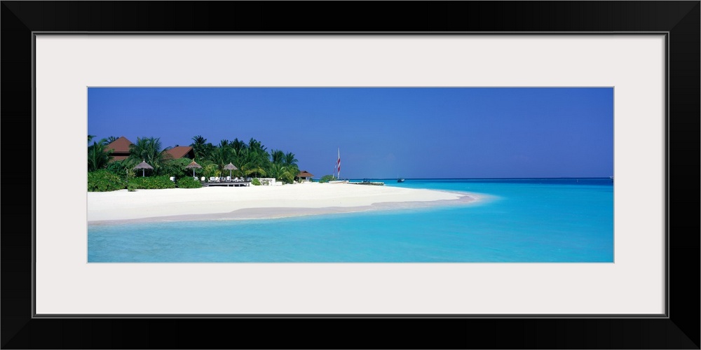 This panoramic photograph is of a beach in Maldives with huts pushed back and surrounded by trees and cool water going aro...