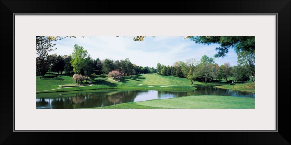 Panoramic photograph of golf course featuring putting green, sand traps, and winding pond.  There are various trees and on...