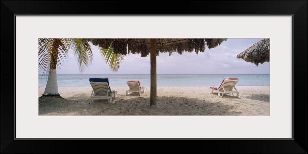 Huge, wide angle photograph of several lounge chairs beneath umbrellas and a palm tree, on 7-Mile Beach in Negril, Jamaica.