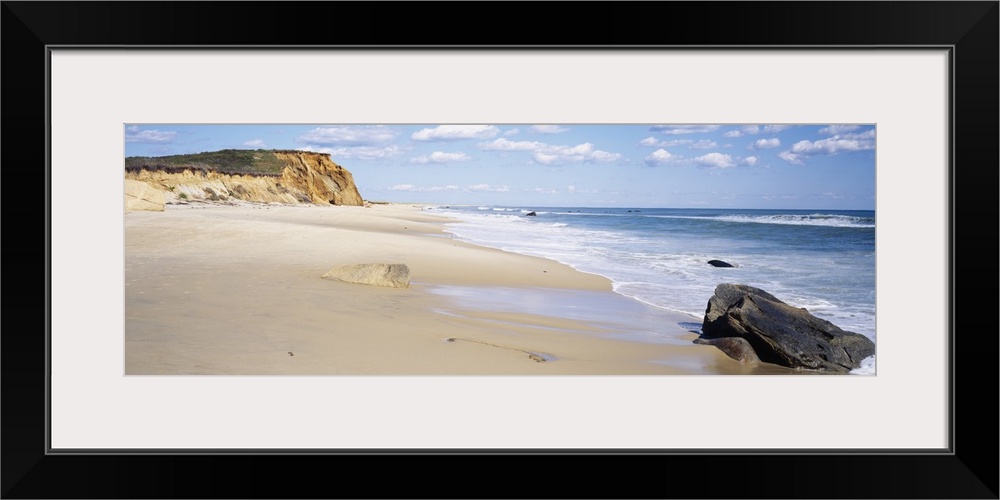 Panoramic photograph of shoreline with surf, large rock, and cliff in the distance under a cloudy sky.