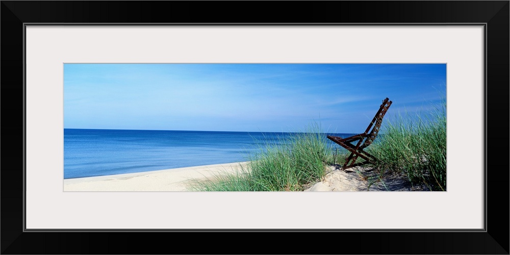 This wall art is a panoramic landscape photograph of a sandy beach with a chair in the dunes overlooking the lake.