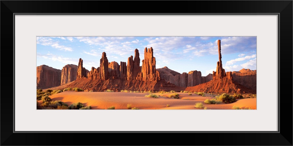 Panoramic photograph composed of a desert landscape filled with sand and small patches of vegetation.  In the background t...