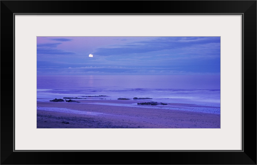 The moon is seen slightly behind a cluster of clouds that hang over the ocean water and photographed in a wide angle view.