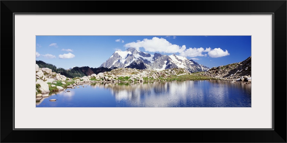 An immense snow covered mountain is pictured in the background from across a large body of water that is surrounded by roc...