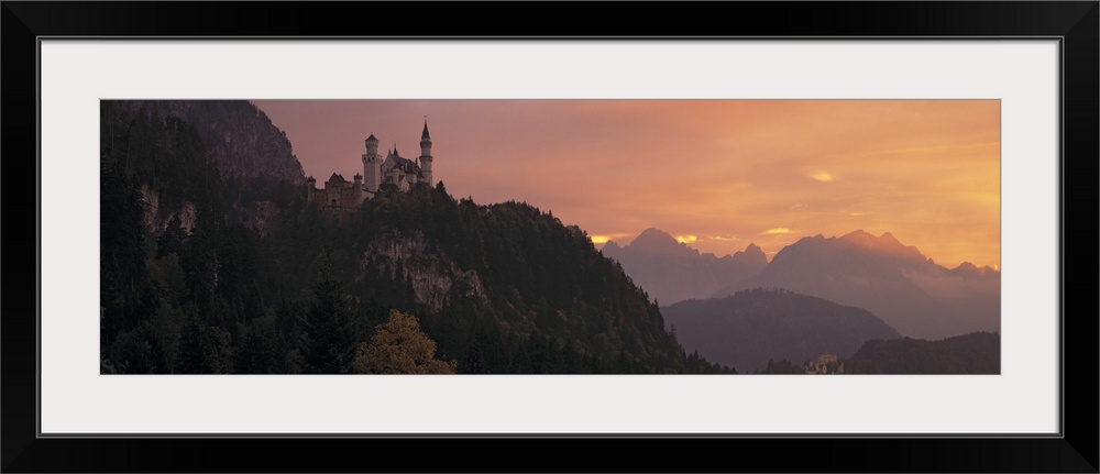 A large panoramic piece of a castle sitting in the mountains in Germany. The sky has warm tones as the sun sets out of view.