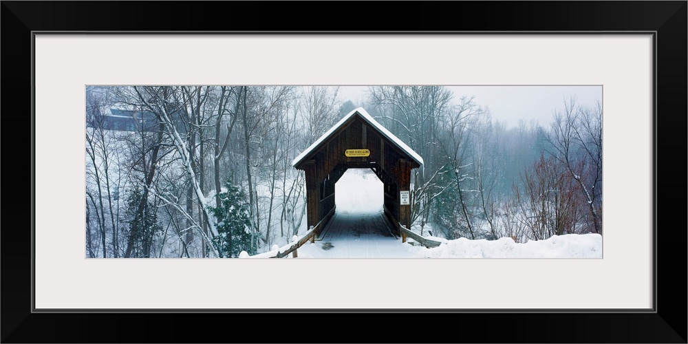 A panoramic photograph of a one lane road through a covered bridge in a winter snowscape.