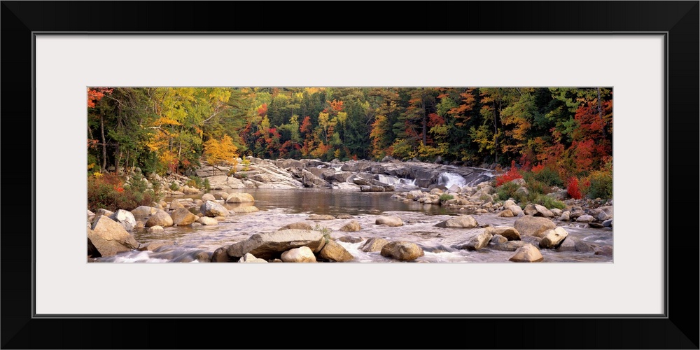 A relaxed panoramic landscape of large boulders in a New England river lined with trees with autumn colors.