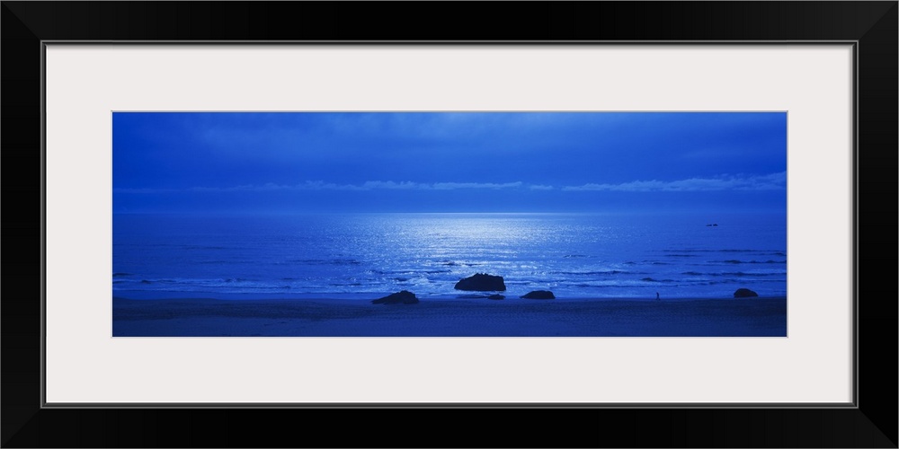 Panoramic photograph of a monotone seascape with the moonlight reflecting off the waves, three solitary rocks sit on the s...