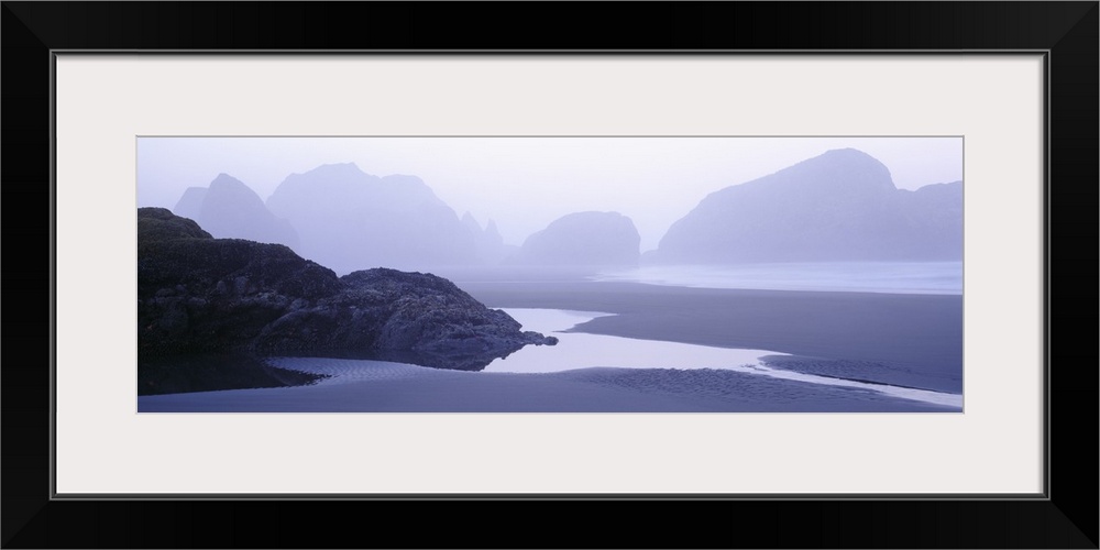 Rock formations in the water are faintly seen behind a layer of fog. Some water puddles on the beach near a smaller rock f...